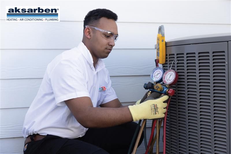 An Aksarben technician in a white shirt and gloves is using a testing tool on a heat pump outside a building.