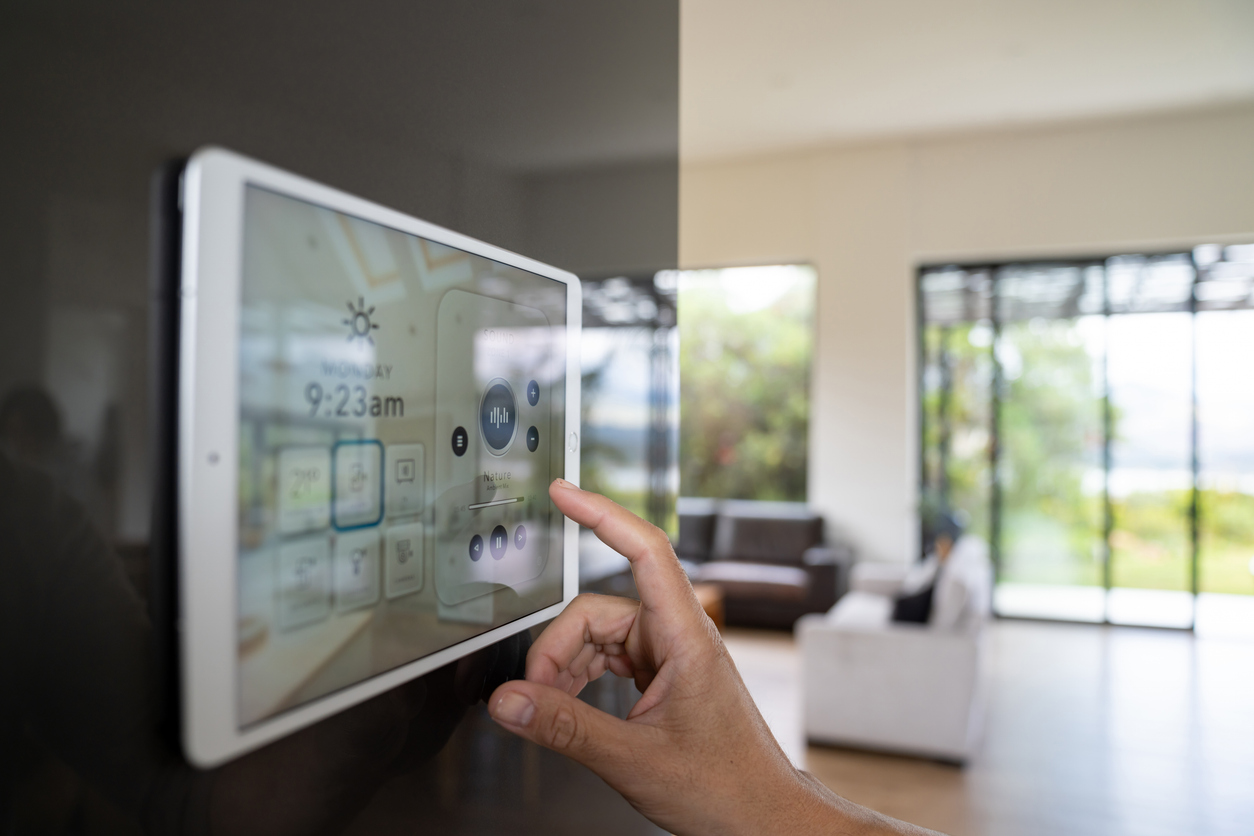 A close-up of a person’s hand tapping a white, wall-mounted touch panel that displays a smart-home interface—showing the day and time, weather icon, temperature settings, and audio controls labeled.